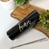 Un elegante termo personalizado liso de 590 ml en color negro, con el nombre Iván escrito en letras blancas, reposa sobre una bandeja de madera. Está rodeado de exuberantes plantas en macetas y viene con una pajita de metal y un cepillo de limpieza. El vaso también muestra el elegante logotipo de Creatiboxs.