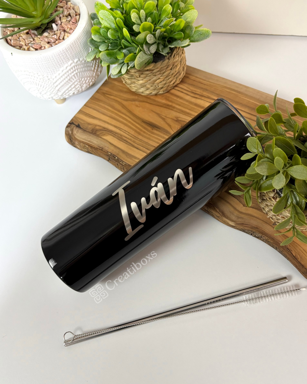 Un elegante termo personalizado liso de 590 ml en color negro, con el nombre Iván escrito en letras blancas, reposa sobre una bandeja de madera. Está rodeado de exuberantes plantas en macetas y viene con una pajita de metal y un cepillo de limpieza. El vaso también muestra el elegante logotipo de Creatiboxs.