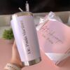 Una persona sostiene un vaso de acero inoxidable con el nombre Lic. Paola Martínez impreso en él. En el fondo hay una caja de regalo rosa con un lazo en el que se puede leer A tu lado siempre, Lic. Pala. Cerca hay una pequeña planta verde.