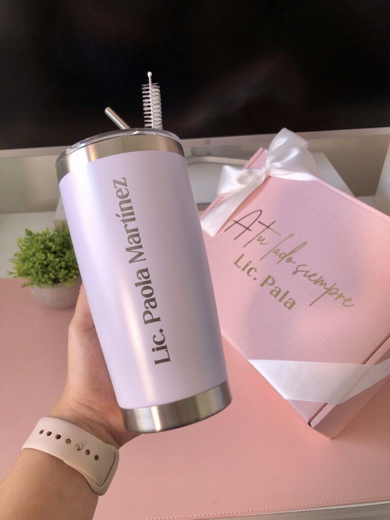 Una persona sostiene un vaso de acero inoxidable con el nombre Lic. Paola Martínez impreso en él. En el fondo hay una caja de regalo rosa con un lazo en el que se puede leer A tu lado siempre, Lic. Pala. Cerca hay una pequeña planta verde.