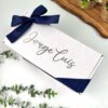 Caja de regalo blanca con nombre en la parte superior, envuelta en liston azul marino y plantas verdes, ideal como regalo para padrino