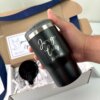 Una mano sostiene un vaso negro grabado con "Jorge Luis 23.08.13" junto a una caja chica personalizada abierta, que presenta el kit de regalo para padrino que incluye termo portalatas 420ml y caja chica personalizada.