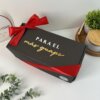 Una caja de regalo negra con una cinta roja y el texto dorado "Para el más guapo" se encuentra sobre una mesa blanca cerca de plantas, presentando el Kit termo portalatas 420ml y caja chica personalizada, un regalo personalizado ideal para cualquier ocasión especial.