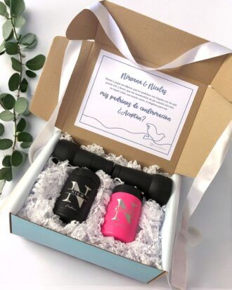 Una caja de regalo abierta contiene dos botellas negras con la palabra Nico y una botella rosa con el mismo diseño. La caja incluye papel decorativo blanco y una tarjeta de invitación con una ilustración de una paloma. Una rama verde con hojas se encuentra al lado de la caja.