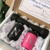 Una caja de regalo que contiene un frasco personalizado de color negro y un artículo personalizado de color rosa, ambos etiquetados con las iniciales N. La caja está llena de papel picado blanco e incluye una tarjeta con texto y una ilustración decorativa de una paloma. Se ve parcialmente una planta cerca.