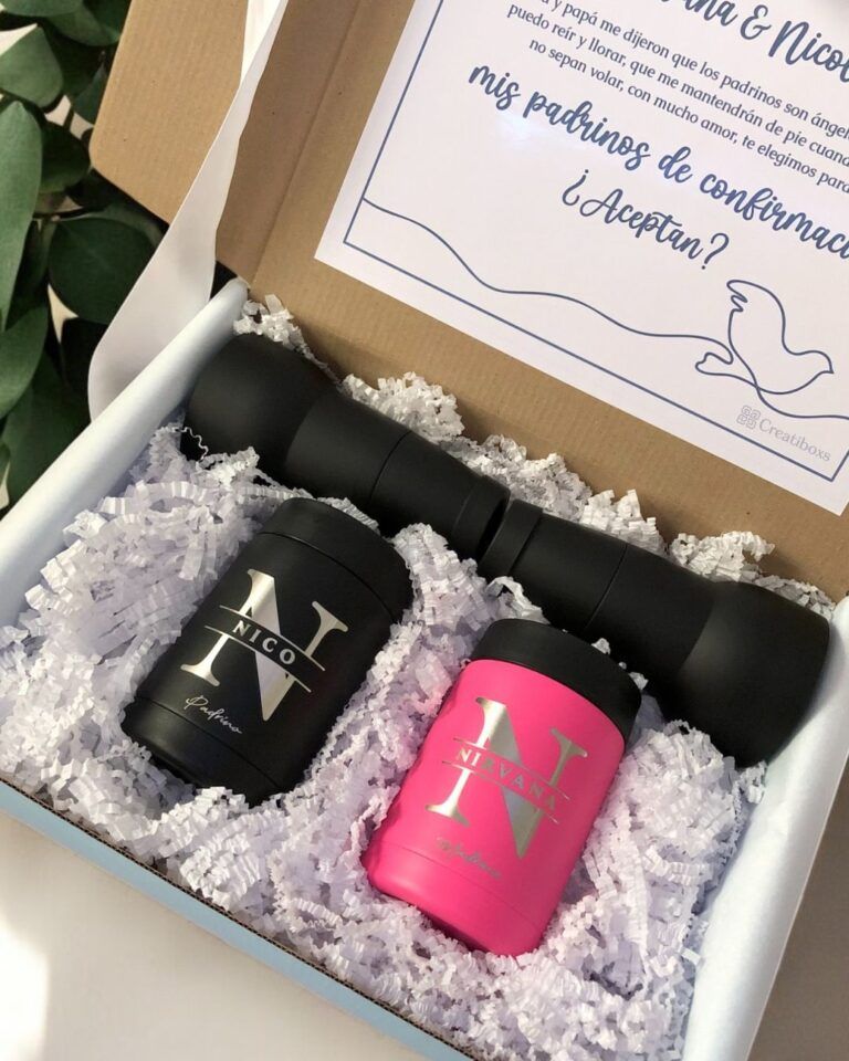 Una caja de regalo que contiene un frasco personalizado de color negro y un artículo personalizado de color rosa, ambos etiquetados con las iniciales N. La caja está llena de papel picado blanco e incluye una tarjeta con texto y una ilustración decorativa de una paloma. Se ve parcialmente una planta cerca.
