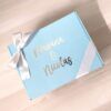 Se muestra una caja de regalo de color azul claro con un lazo blanco. La caja tiene una etiqueta con la palabra Nirvana & Nicolás en letras metálicas y blancas. El nombre de la marca Creatiboxs se encuentra en la esquina inferior. La caja está colocada sobre una superficie de madera.