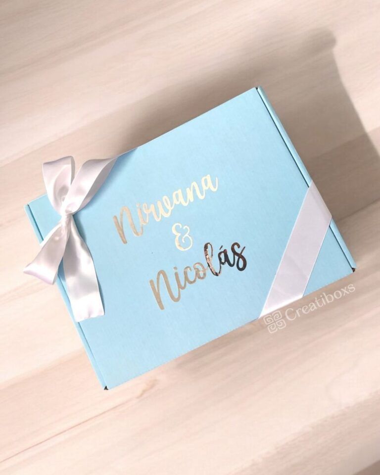 Se muestra una caja de regalo de color azul claro con un lazo blanco. La caja tiene una etiqueta con la palabra Nirvana & Nicolás en letras metálicas y blancas. El nombre de la marca Creatiboxs se encuentra en la esquina inferior. La caja está colocada sobre una superficie de madera.