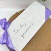 Una caja rectangular con una cinta morada sobre una estera tejida. La caja tiene un texto en español que dice Tía, hay algo que quiero decirte. En el fondo, hay suculentas verdes.