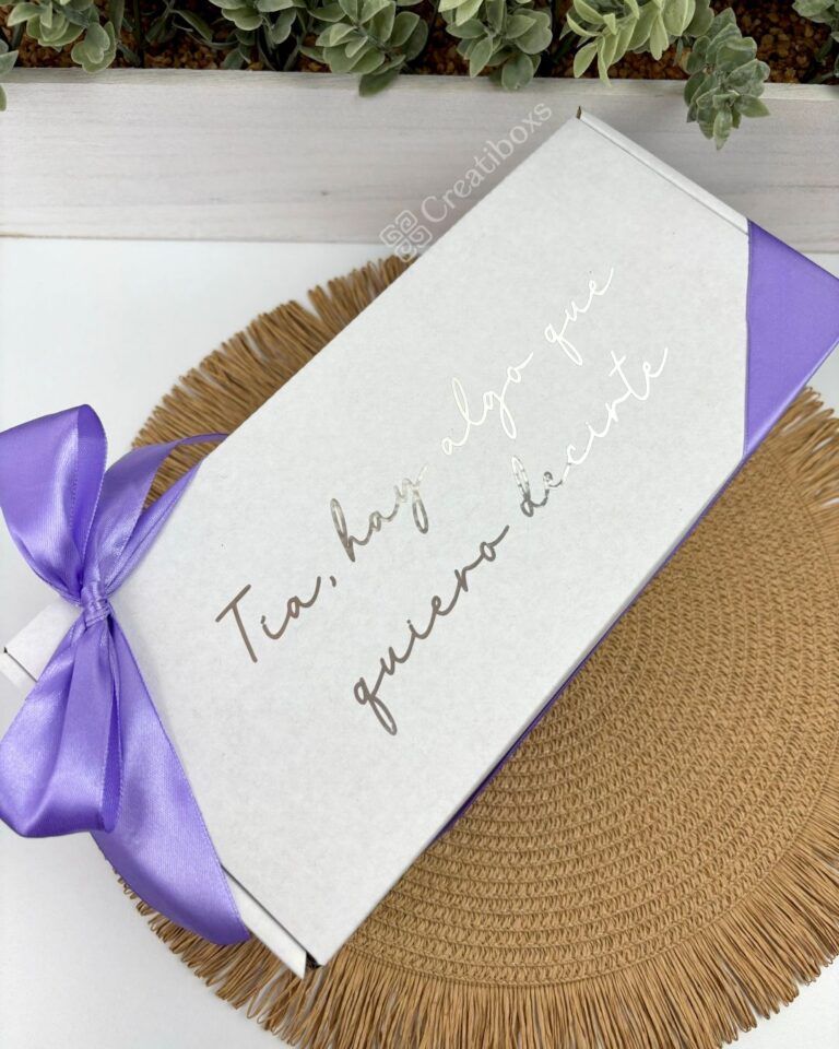 Una caja rectangular con una cinta morada sobre una estera tejida. La caja tiene un texto en español que dice Tía, hay algo que quiero decirte. En el fondo, hay suculentas verdes.