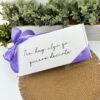 Una caja rectangular blanca con el texto Tía, hay algo que quiero decirte, atada con una cinta morada. La caja está colocada sobre un mantel circular tejido. Detrás de ella hay una hilera de pequeñas plantas verdes en una jardinera.