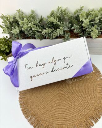 Una caja rectangular blanca con el texto Tía, hay algo que quiero decirte, atada con una cinta morada. La caja está colocada sobre un mantel circular tejido. Detrás de ella hay una hilera de pequeñas plantas verdes en una jardinera.