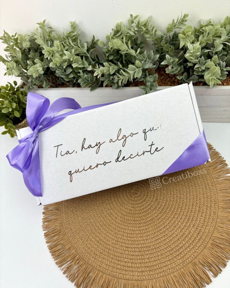 Una caja rectangular blanca con el texto Tía, hay algo que quiero decirte, atada con una cinta morada. La caja está colocada sobre un mantel circular tejido. Detrás de ella hay una hilera de pequeñas plantas verdes en una jardinera.