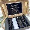 Una caja de regalo que contiene dos vasos, uno blanco y otro negro, colocados sobre papel arrugado negro. También hay dos pajitas de metal y un cepillo de limpieza. La tapa tiene una tarjeta con un texto en español que expresa gratitud y una invitación. Una cinta dorada decora la caja.