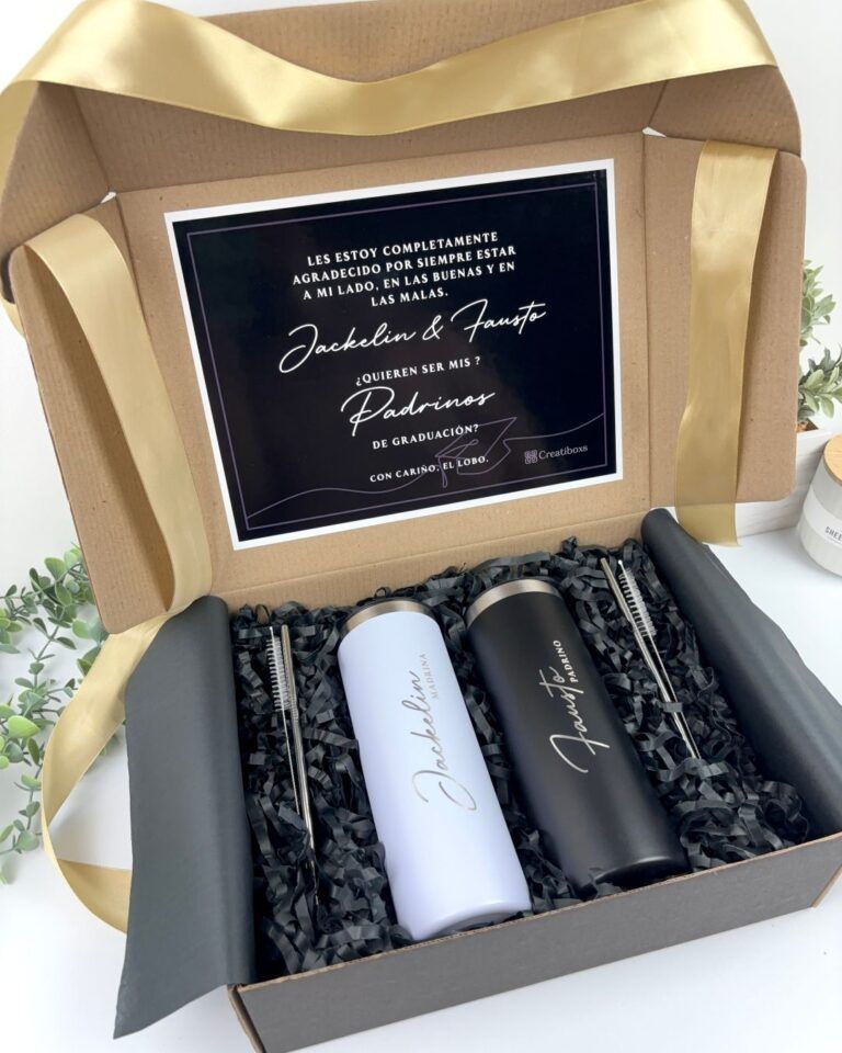 Una caja de regalo que contiene dos vasos, uno blanco y otro negro, colocados sobre papel arrugado negro. También hay dos pajitas de metal y un cepillo de limpieza. La tapa tiene una tarjeta con un texto en español que expresa gratitud y una invitación. Una cinta dorada decora la caja.