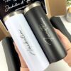 Dos vasos térmicos, uno blanco y otro negro, se sostienen en las manos. Cada vaso presenta una elegante escritura manuscrita. Parecen ser parte de un paquete de regalo, con una caja de cartón parcialmente visible en el fondo.