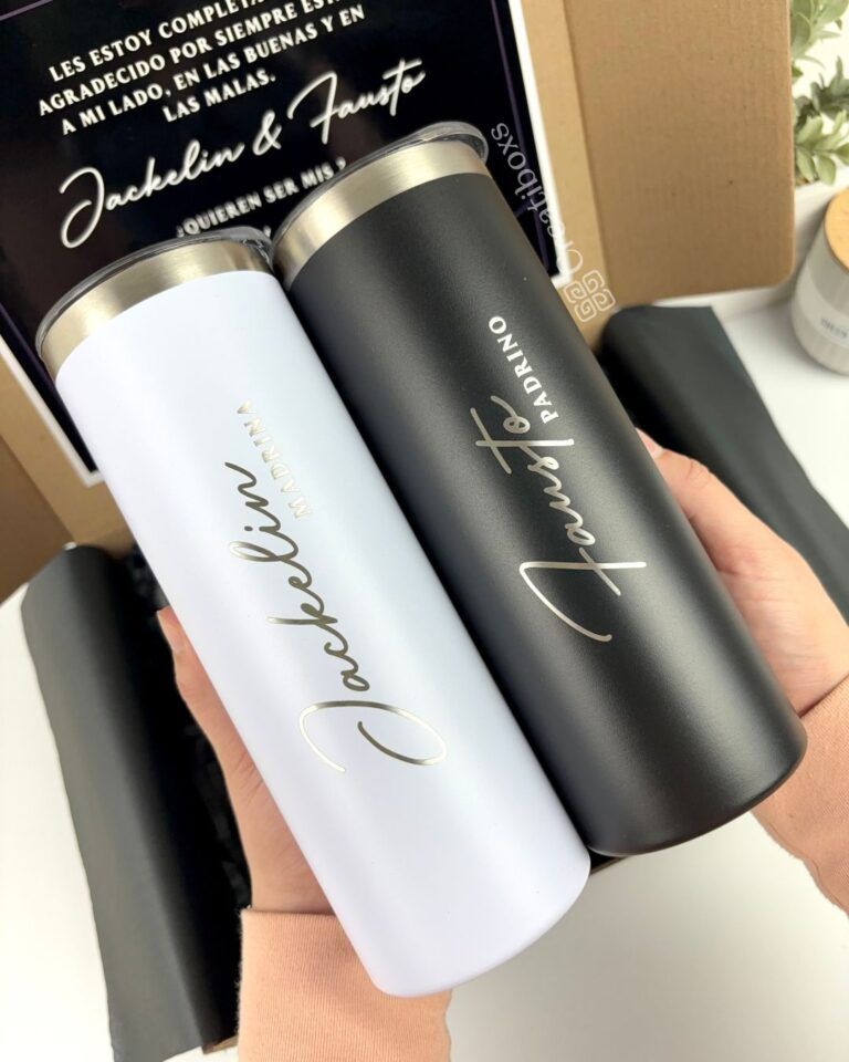 Dos vasos térmicos, uno blanco y otro negro, se sostienen en las manos. Cada vaso presenta una elegante escritura manuscrita. Parecen ser parte de un paquete de regalo, con una caja de cartón parcialmente visible en el fondo.