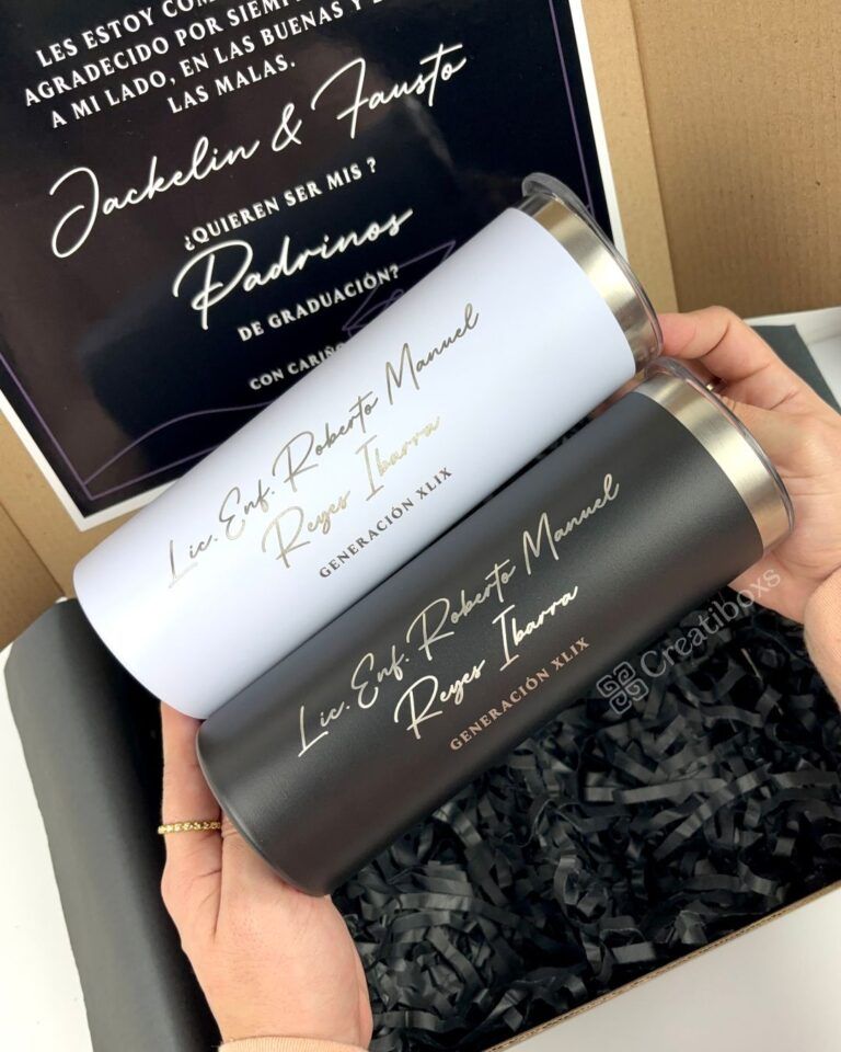 Dos vasos, uno blanco y otro negro, con un elegante texto escrito a mano, se sostienen sobre una caja de regalo negra con papel picado. En el fondo se ve parcialmente una tarjeta impresa con un diseño decorativo.