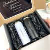 Una caja de regalo contiene dos tazas de viaje, una blanca con la palabra Jackelin impresa en negro y otra negra con la palabra Fausto. Están envueltas en papel arrugado negro. Una nota en la tapa de la caja incluye el texto padrinos de graduación y ¿Quieren ser mis?.