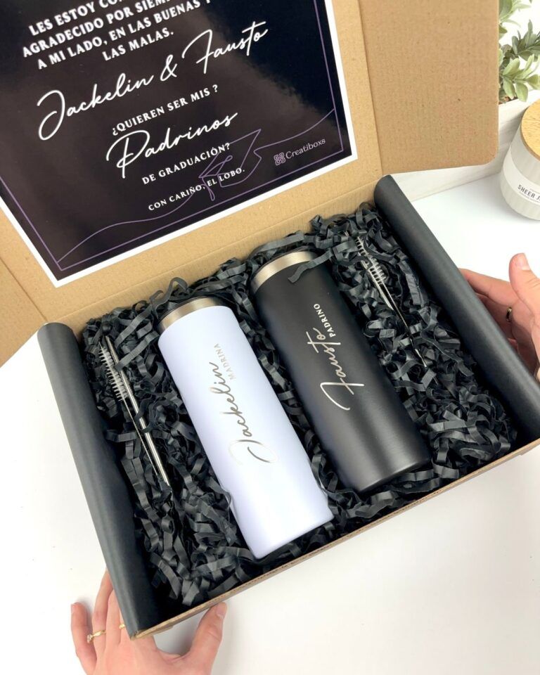 Una caja de regalo contiene dos tazas de viaje, una blanca con la palabra Jackelin impresa en negro y otra negra con la palabra Fausto. Están envueltas en papel arrugado negro. Una nota en la tapa de la caja incluye el texto padrinos de graduación y ¿Quieren ser mis?.