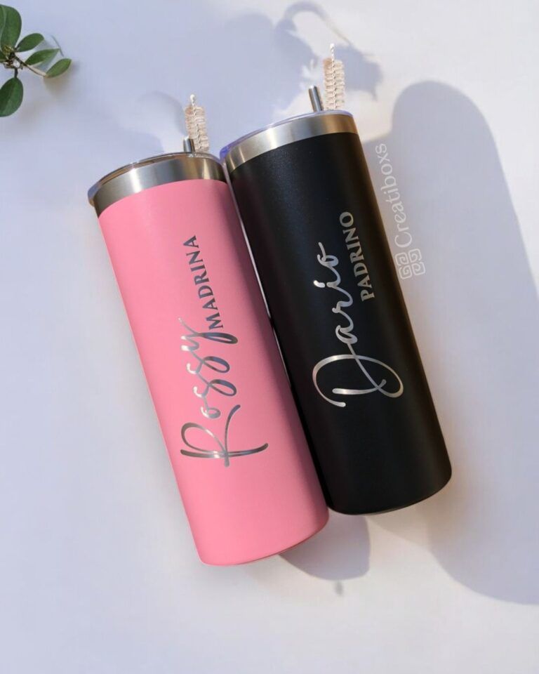 Dos vasos altos se colocan uno al lado del otro sobre una superficie plana. El vaso rosa tiene escrito Rosa Madrina y el vaso negro tiene escrito Dario Padrino. Ambos tienen tapas de metal con pajitas. Se ve una pequeña planta en la esquina.