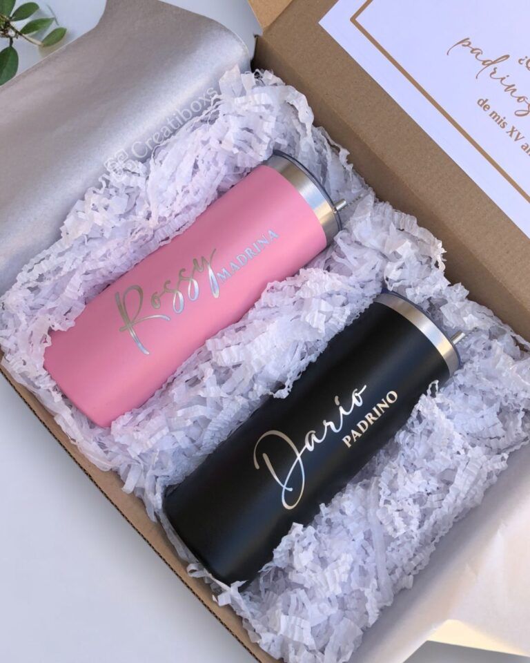 Una caja contiene dos vasos colocados en papel triturado blanco. El vaso izquierdo es rosa y tiene escrito Rossey Madrina y el vaso derecho es negro y tiene escrito Dario Padrino. Están rodeados por un forro de papel de seda beige.
