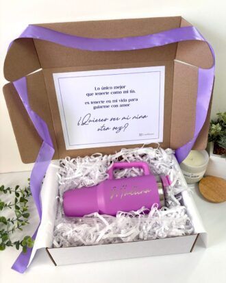Una caja de regalo contiene un vaso rosa con la palabra Madrina escrita en él, rodeado de material de embalaje blanco. La tapa es rosa y una cinta lavanda adorna la caja. Una nota impresa en español es visible sobre el vaso.