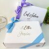 Se muestran dos cajas de regalo blancas con lazos de cinta de color pastel. La caja superior tiene un lazo morado y dice Enf. Silvia, mientras que la caja inferior tiene un lazo azul y dice Enf. Elba García. Una pequeña planta es parcialmente visible junto a las cajas.