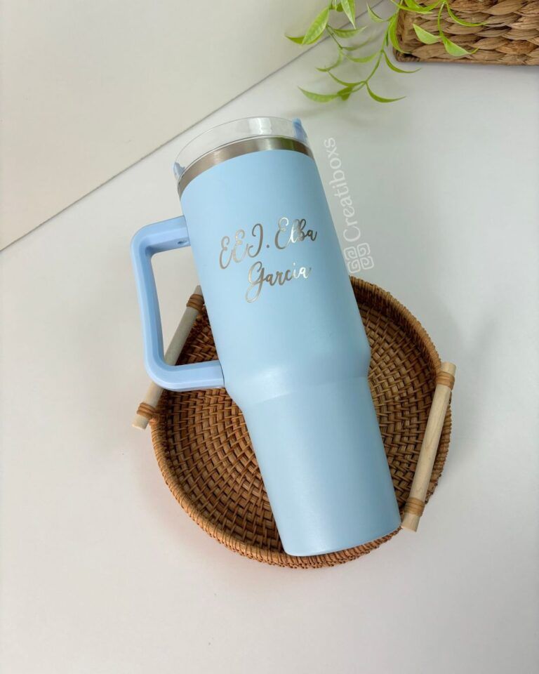 Sobre una bandeja redonda de mimbre se encuentra un vaso azul claro con asa. El nombre E.J. Ella García está grabado en el vaso. En la esquina superior derecha se ve una pequeña porción de una planta verde.