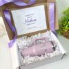 Una caja de regalo contiene un vaso térmico de color lavanda con el texto Madrina 2023 y una tarjeta que dice ¿Le gustaría ser nuestra Madrina de generación?. La caja está forrada con papel arrugado blanco y decorada con una cinta morada.