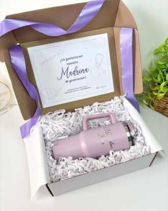 Una caja de regalo contiene un vaso térmico de color lavanda con el texto Madrina 2023 y una tarjeta que dice ¿Le gustaría ser nuestra Madrina de generación?. La caja está forrada con papel arrugado blanco y decorada con una cinta morada.