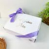 Se muestra una caja de regalo blanca con una cinta violeta. La caja tiene impreso el texto Eab, Silvia, Jana. Junto a la caja, sobre una superficie blanca, hay una estera circular tejida y una pequeña planta verde en maceta.