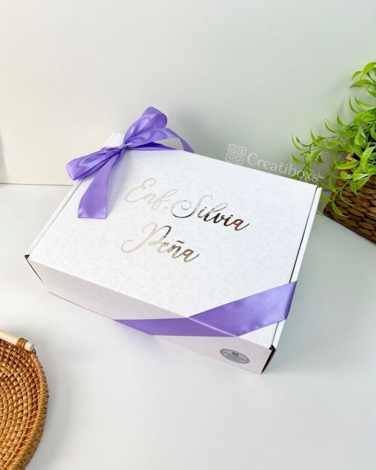 Se muestra una caja de regalo blanca con una cinta violeta. La caja tiene impreso el texto Eab, Silvia, Jana. Junto a la caja, sobre una superficie blanca, hay una estera circular tejida y una pequeña planta verde en maceta.