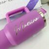 Vaso térmico morado con la palabra Madrina escrita en cursiva en el lateral. El vaso tiene asa y tapón de acero inoxidable. Está colocado delante de una caja blanca con texto parcialmente visible.