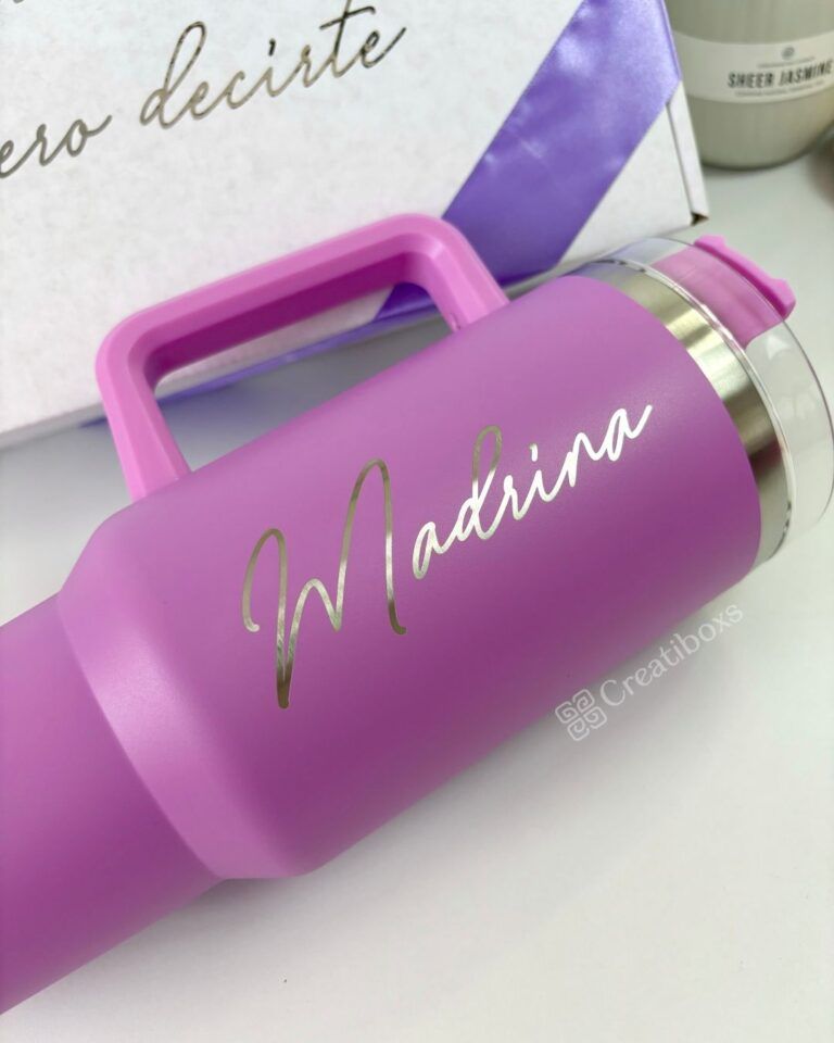 Vaso térmico morado con la palabra Madrina escrita en cursiva en el lateral. El vaso tiene asa y tapón de acero inoxidable. Está colocado delante de una caja blanca con texto parcialmente visible.