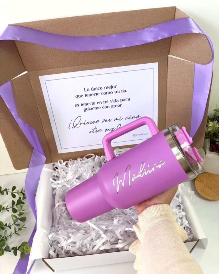 Una mano sostiene un vaso rosa con la palabra Madrina escrita. El vaso está dentro de una caja abierta con cintas rosas y papel arrugado blanco. Una tarjeta en el interior dice: Lo único mejor que tenerte como mi tía, es tenerte en mi vida para guiarme con amor. ¿Quieres ser mi madrina otra vez?.