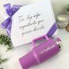 Una caja de regalo blanca con un lazo violeta y el texto en español Tía hay algo importante que quiero decirte. Delante de la caja hay una taza de viaje violeta con la palabra Madrina impresa. Cerca hay una vela y hojas verdes.