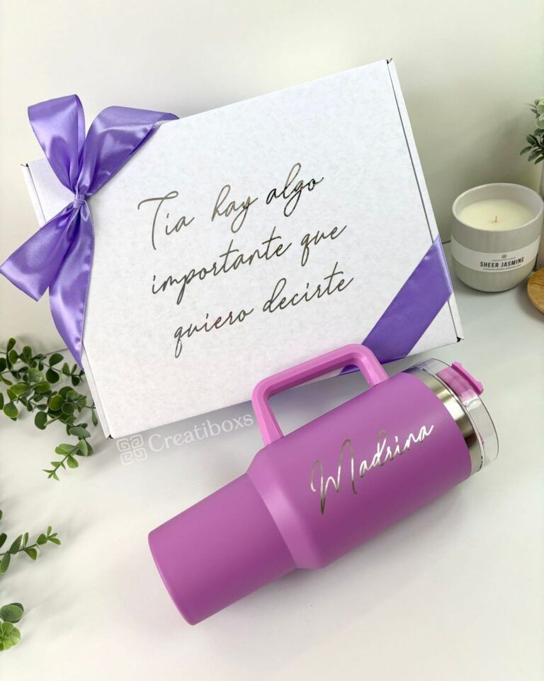 Una caja de regalo blanca con un lazo violeta y el texto en español Tía hay algo importante que quiero decirte. Delante de la caja hay una taza de viaje violeta con la palabra Madrina impresa. Cerca hay una vela y hojas verdes.