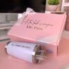 Una caja de regalo de color rosa con la inscripción A tu lado siempre Lic. Pala en la tapa, acompañada de un vaso blanco con la etiqueta Lic. Paola Martínez. La caja está envuelta con un lazo blanco. En el fondo, sobre un escritorio, hay una pequeña planta.