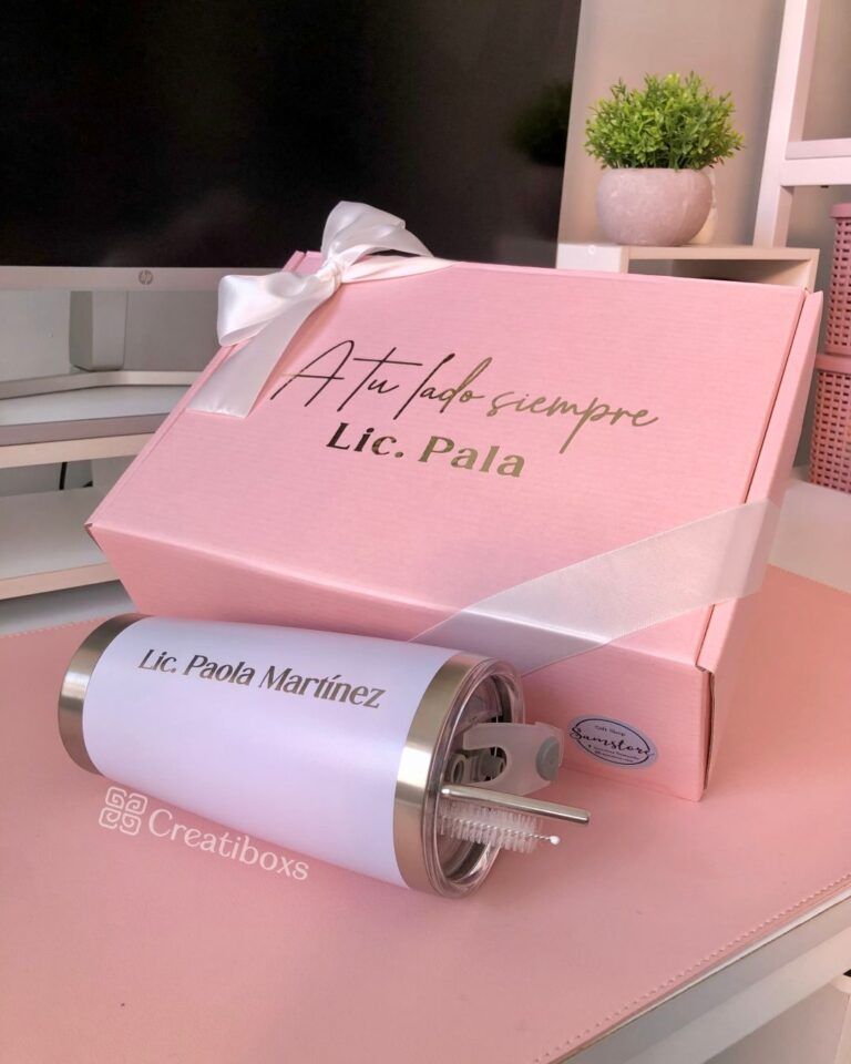 Una caja de regalo de color rosa con la inscripción A tu lado siempre Lic. Pala en la tapa, acompañada de un vaso blanco con la etiqueta Lic. Paola Martínez. La caja está envuelta con un lazo blanco. En el fondo, sobre un escritorio, hay una pequeña planta.