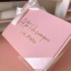 Sobre una superficie se coloca una caja de regalo de color rosa con un lazo blanco. La caja tiene un texto dorado que dice A tu lado siempre, Lic. Pala. El logotipo de Creatiboxes se encuentra en la esquina inferior izquierda de la caja. En el fondo se ve parte de un monitor y un estante.