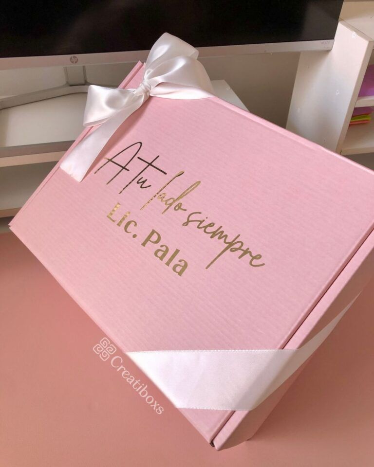 Sobre una superficie se coloca una caja de regalo de color rosa con un lazo blanco. La caja tiene un texto dorado que dice A tu lado siempre, Lic. Pala. El logotipo de Creatiboxes se encuentra en la esquina inferior izquierda de la caja. En el fondo se ve parte de un monitor y un estante.