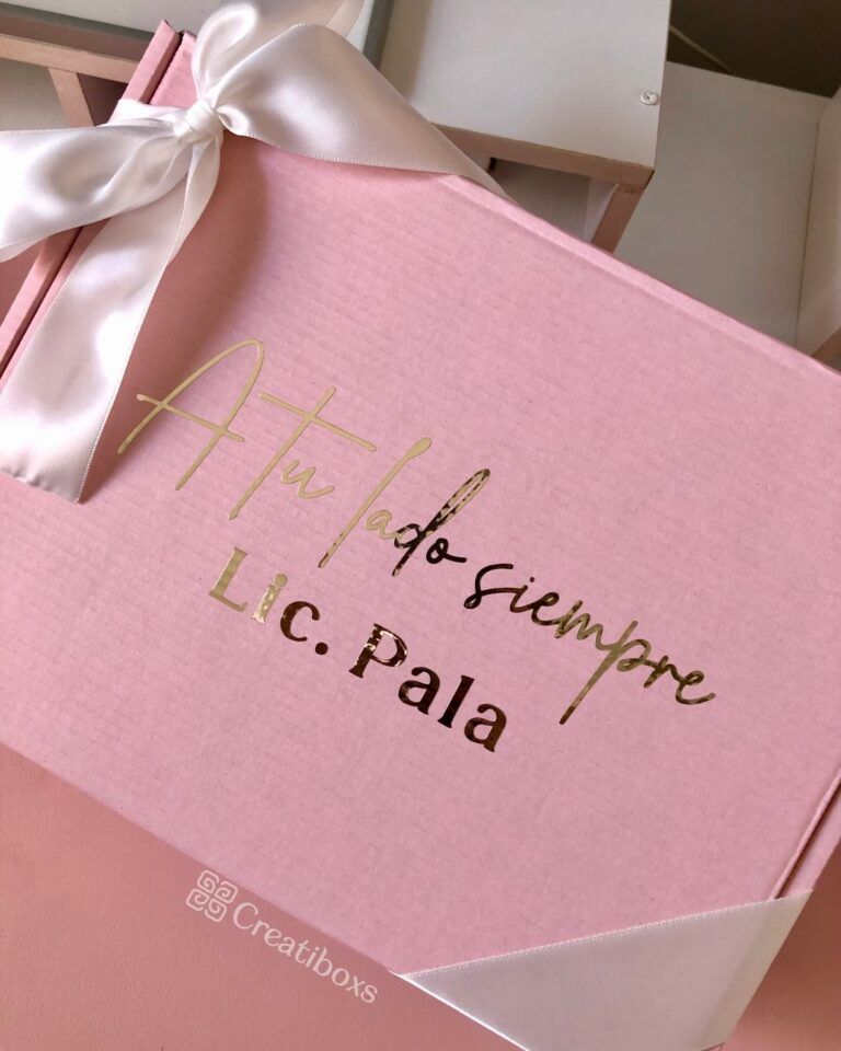 Una caja de regalo rosa con una cinta blanca que incluye un texto dorado que dice A tu lado siempre y Lic. Pala. El logotipo Creatiboxs está impreso en blanco en la esquina inferior de la caja.