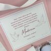 Una caja de regalo rosa y blanca que contiene una tarjeta con texto en español dirigida a una Madrina. La tarjeta tiene ilustraciones de palomas y viene acompañada de papel de embalaje triturado y cinta.