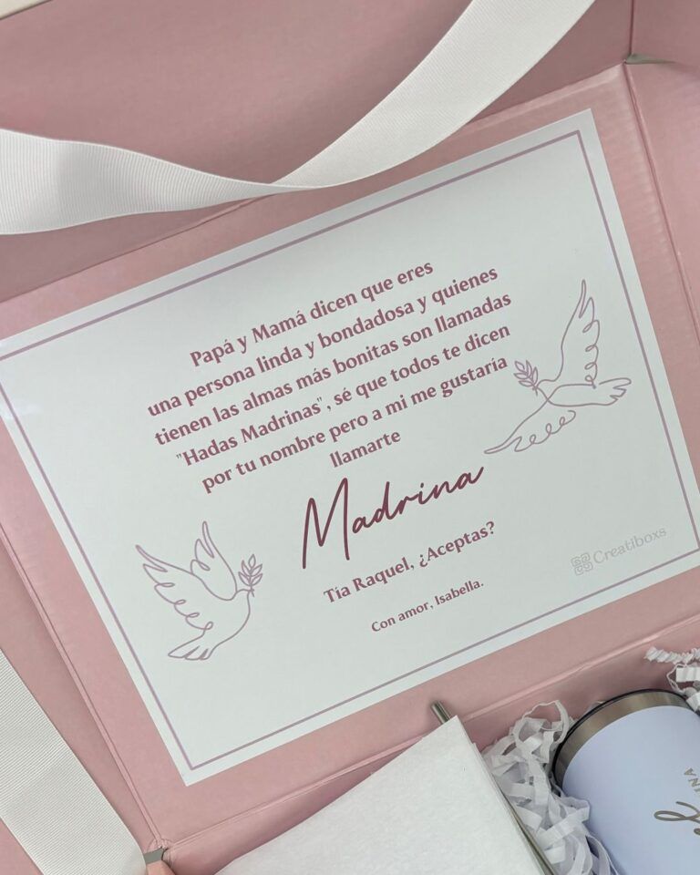 Una caja de regalo rosa y blanca que contiene una tarjeta con texto en español dirigida a una Madrina. La tarjeta tiene ilustraciones de palomas y viene acompañada de papel de embalaje triturado y cinta.