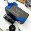 Una caja de regalo negra con un lazo azul se encuentra junto a un vaso negro personalizado con la palabra Ken escrita en él. La caja dice Tío, hay algo que quiero decirte en letras blancas. Se ven pequeñas plantas decorativas y un cartel de madera en el fondo.