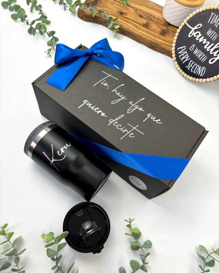 Una caja de regalo negra con un lazo azul se encuentra junto a un vaso negro personalizado con la palabra Ken escrita en él. La caja dice Tío, hay algo que quiero decirte en letras blancas. Se ven pequeñas plantas decorativas y un cartel de madera en el fondo.