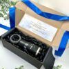 Una botella negra con aislamiento y un logotipo se encuentra dentro de una caja de cartón abierta forrada con papel negro triturado. Se incluye una nota doblada y una cinta de raso azul. La caja está rodeada de hojas de eucalipto.