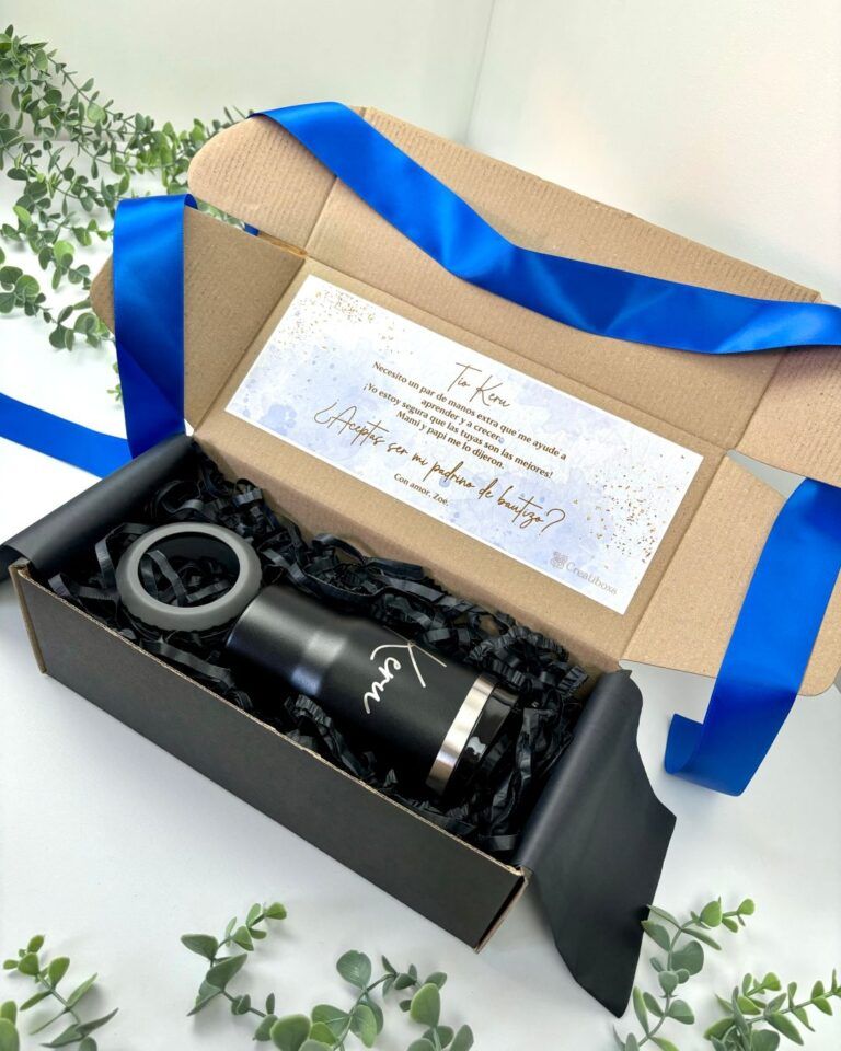 Una botella negra con aislamiento y un logotipo se encuentra dentro de una caja de cartón abierta forrada con papel negro triturado. Se incluye una nota doblada y una cinta de raso azul. La caja está rodeada de hojas de eucalipto.