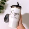 Una mano sostiene una taza de viaje blanca con el nombre Sergio Padrino impreso en letras mayúsculas y cursivas. La taza tiene una tapa negra y, al fondo, una planta con hojas verdes.